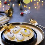Luftige Schneenockerl mit Vanillesauce auf einem dunklen Teller mit einem gold-farbigen Unterteller. Dahinter weihnachtliches Dekor