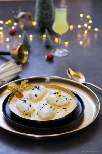 Luftige Schneenockerl mit Vanillesauce auf einem dunklen Teller mit einem gold-farbigen Unterteller. Dahinter weihnachtliches Dekor